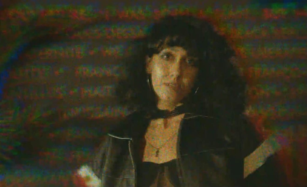 A woman with curly hair wearing a black jacket and choker, standing in front of a colorful, distorted background with text patterns.