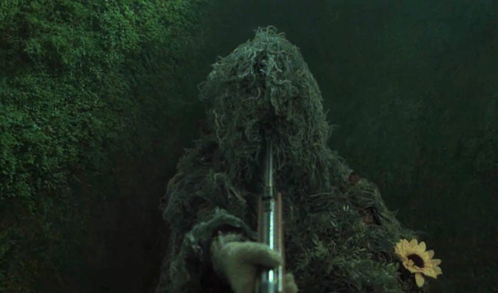 A person camouflaged in foliage aiming a rifle, with a sunflower visible on their outfit, set against a green, forest-like background.