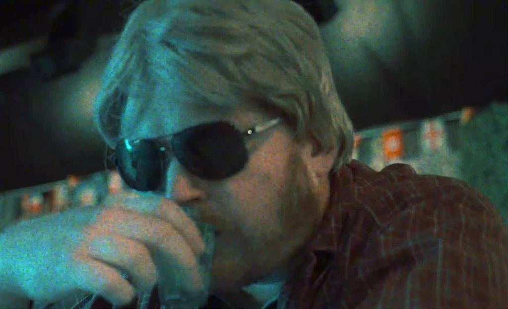 Man with blond hair and sunglasses sipping a drink in a dimly lit bar.
