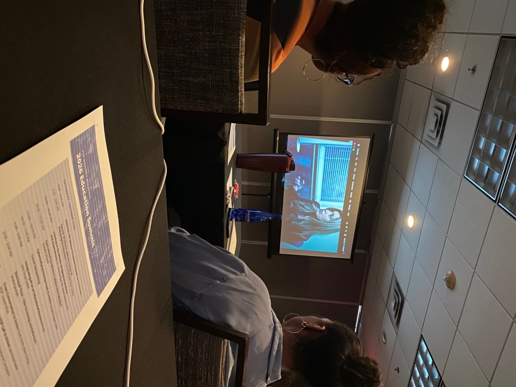 Attendees at a conference viewing a presentation on a screen featuring a woman speaking.