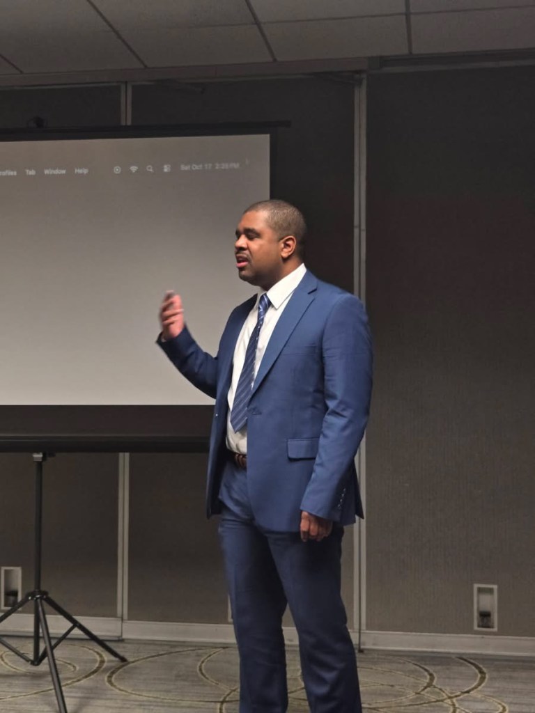 A man in a blue suit standing while presenting in front of a screen in a conference room.