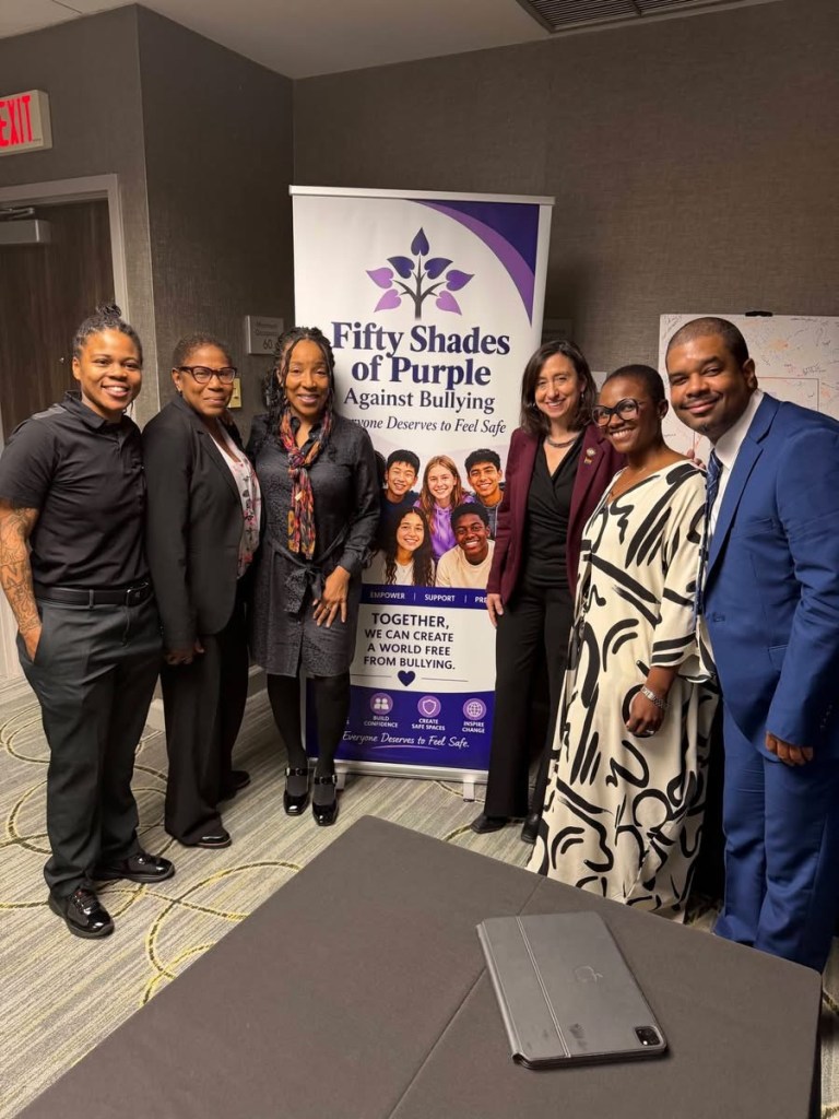 A group of six individuals posing for a photo in front of a banner for 'Fifty Shades of Purple Against Bullying', which promotes safety and anti-bullying awareness.