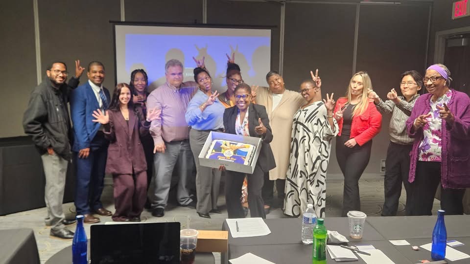 A group of diverse individuals poses for a photo in a conference room, some holding up peace signs. They appear to be celebrating an achievement, with a projector screen in the background.