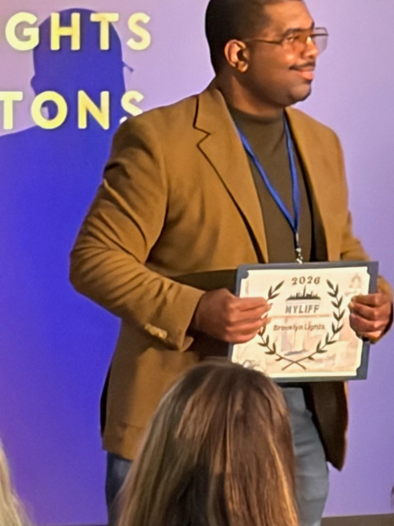 A man wearing a brown blazer holds an award certificate with the year 2020 and the name 'Breawnlyn Liggins' while standing in front of a backdrop with the words 'Insignts' and 'Connections'.
